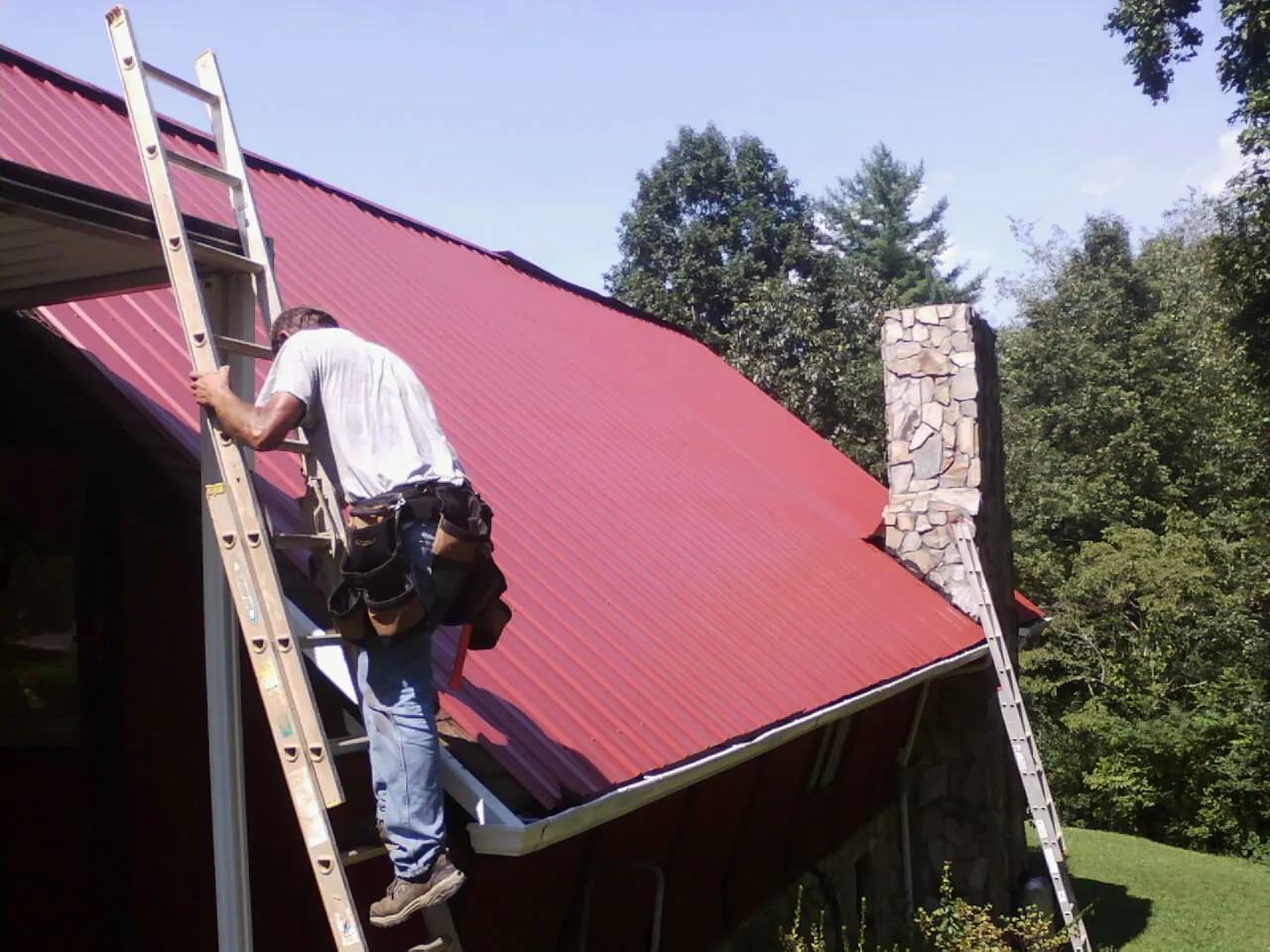 Roofer on a ladder beside a red metal roof and stone chimney — Soffit & Fascia Repair by Roofing Brothers in Pine Valley